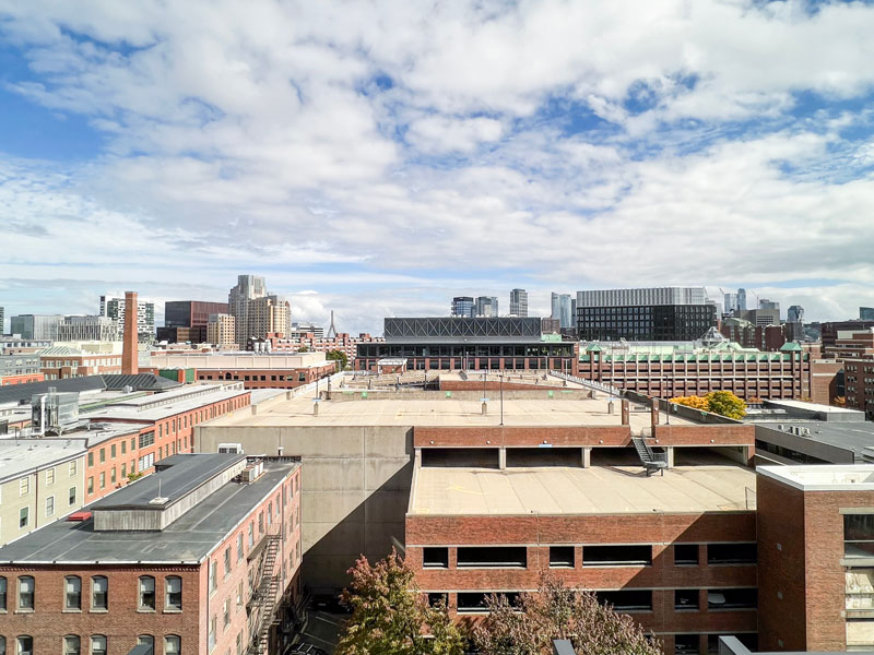 East-facing urban landscape showing rooftops and distant skyscrapers under blue skies and fluffy clouds