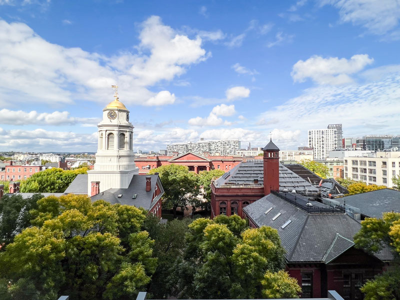 View of historic clock tower in Cambridge surrounded by trees, modern buildings, and blue sky
